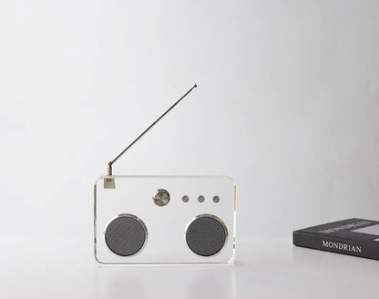 A small rectangular portable radio with a transparent acrylic body, two round black speaker grilles, a silver tuning knob, and an extended black antenna, shown on a white surface against a light gray background. A black book with the word 'MONDRIAN' is partially visible at the right edge.