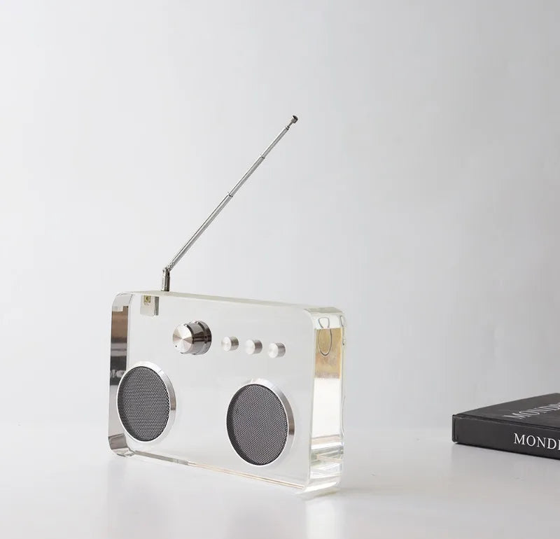 A modern rectangular portable radio made of transparent acrylic with a silver metal antenna, speaker grilles, and control dial, shown on a white surface against a light gray background.