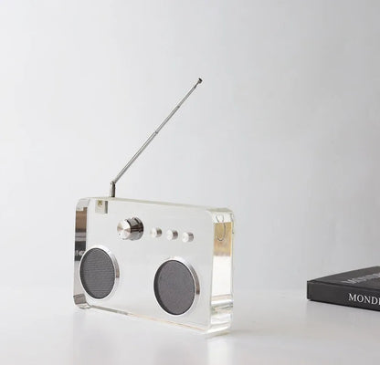 A modern rectangular portable radio made of transparent acrylic with a silver metal antenna, speaker grilles, and control dial, shown on a white surface against a light gray background.