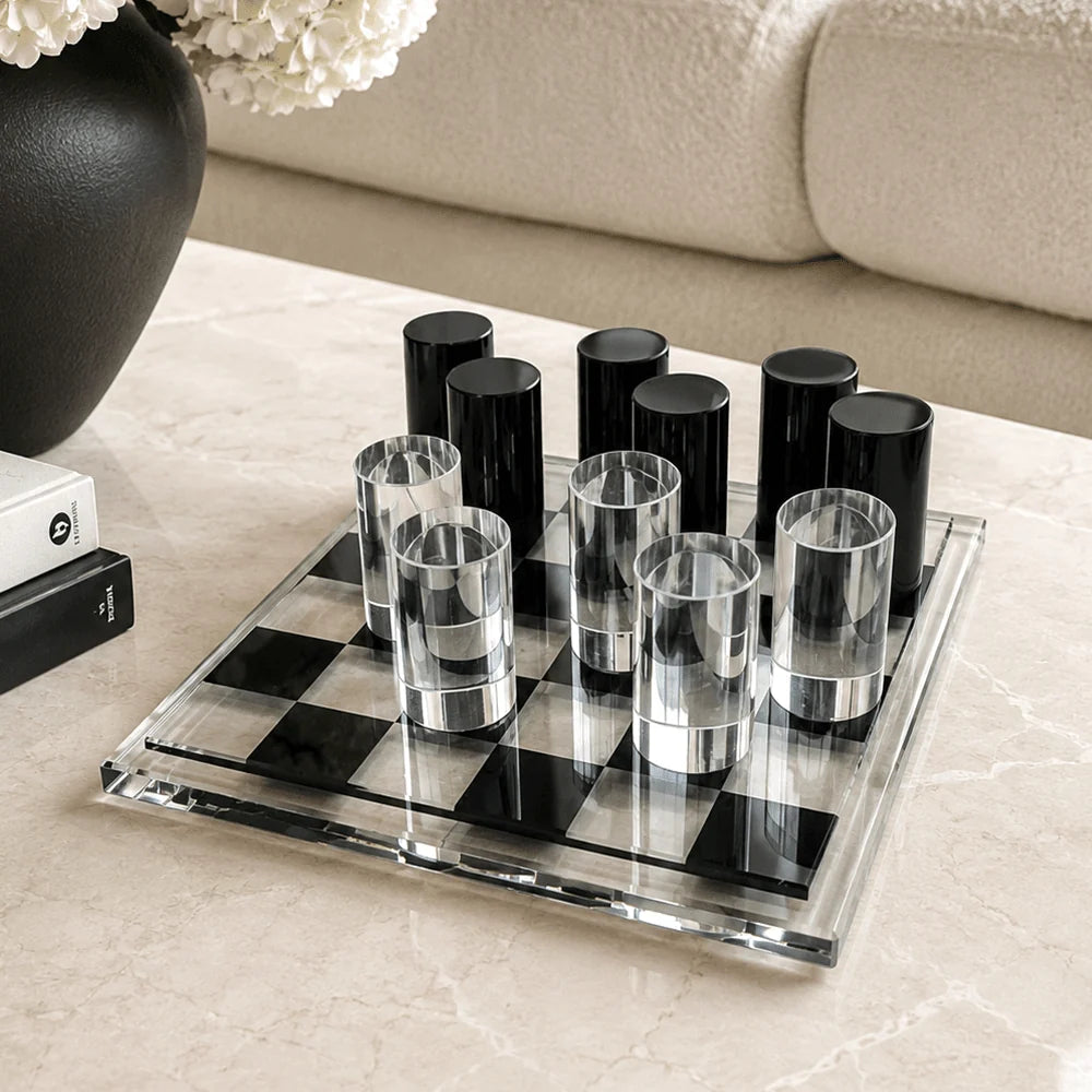 A clear glass checkered game board with black-and-clear cups arranged on it, set on a light marble tabletop in a living room near a sofa and a dark vase.