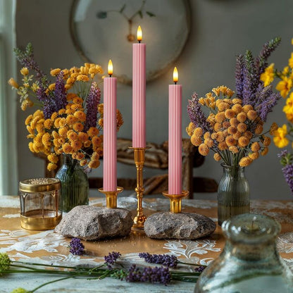 Three pink ribbed flameless taper candles glowing in gold candlestick holders, styled with wildflowers and lavender