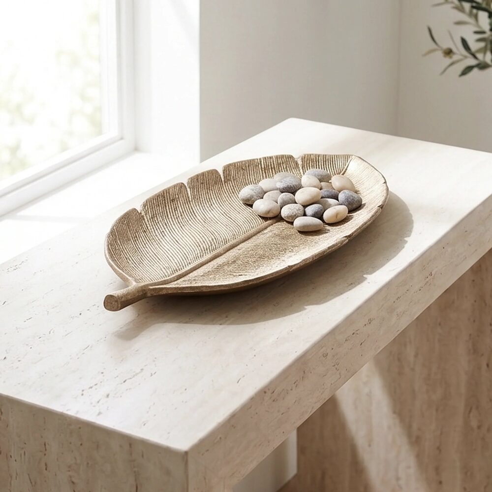 Gold leaf-shaped decorative tray holding small white and grey pebbles, on a light stone windowsill