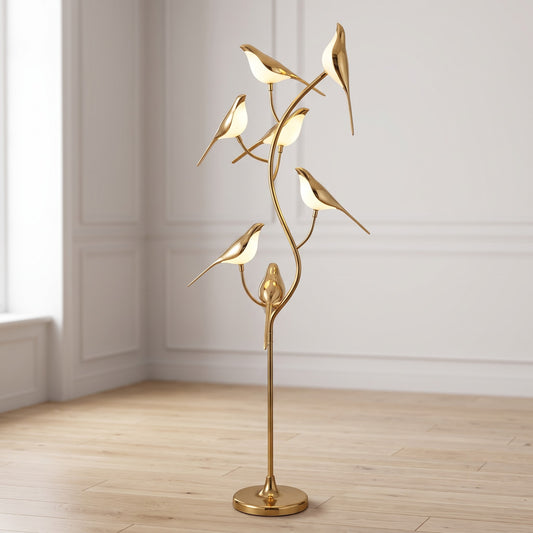 A modern floor lamp shaped like a gold tree branch with several small bird figures and glowing white oval lights, shown in a bright room with pale walls and wood flooring.