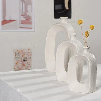 Three modern white ceramic arch-shaped vases of different sizes arranged on a white table, with two vases containing dried yellow flowers. The background is a softly blurred light gray wall with framed art.