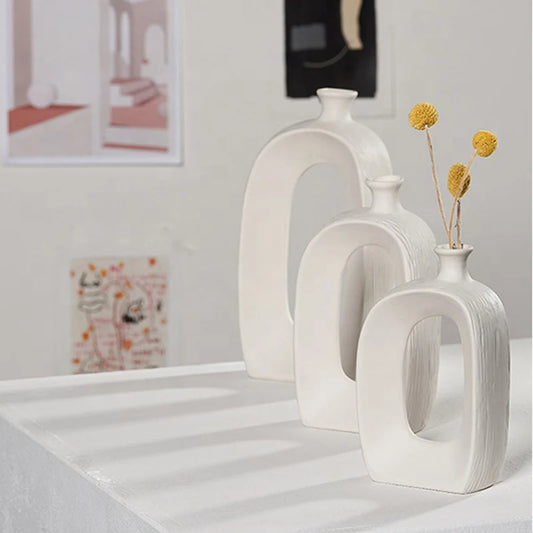 Three modern white ceramic arch-shaped vases of different sizes arranged on a white table, with two vases containing dried yellow flowers. The background is a softly blurred light gray wall with framed art.